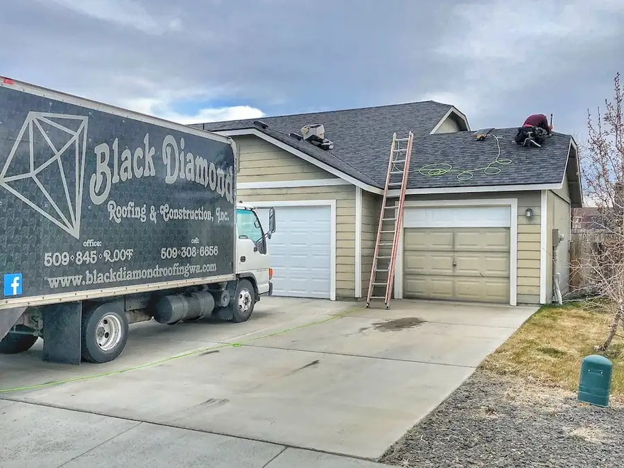 Black Diamond's black box truck in a customer's driveway, ladder up, a roofer working on the new roof